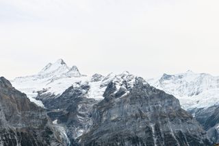 Snowy peaks of a mountain range against a bright and clouded sky. There is a strong contrast between the dark rock structure of the peaks and the lighter snow and sky.
