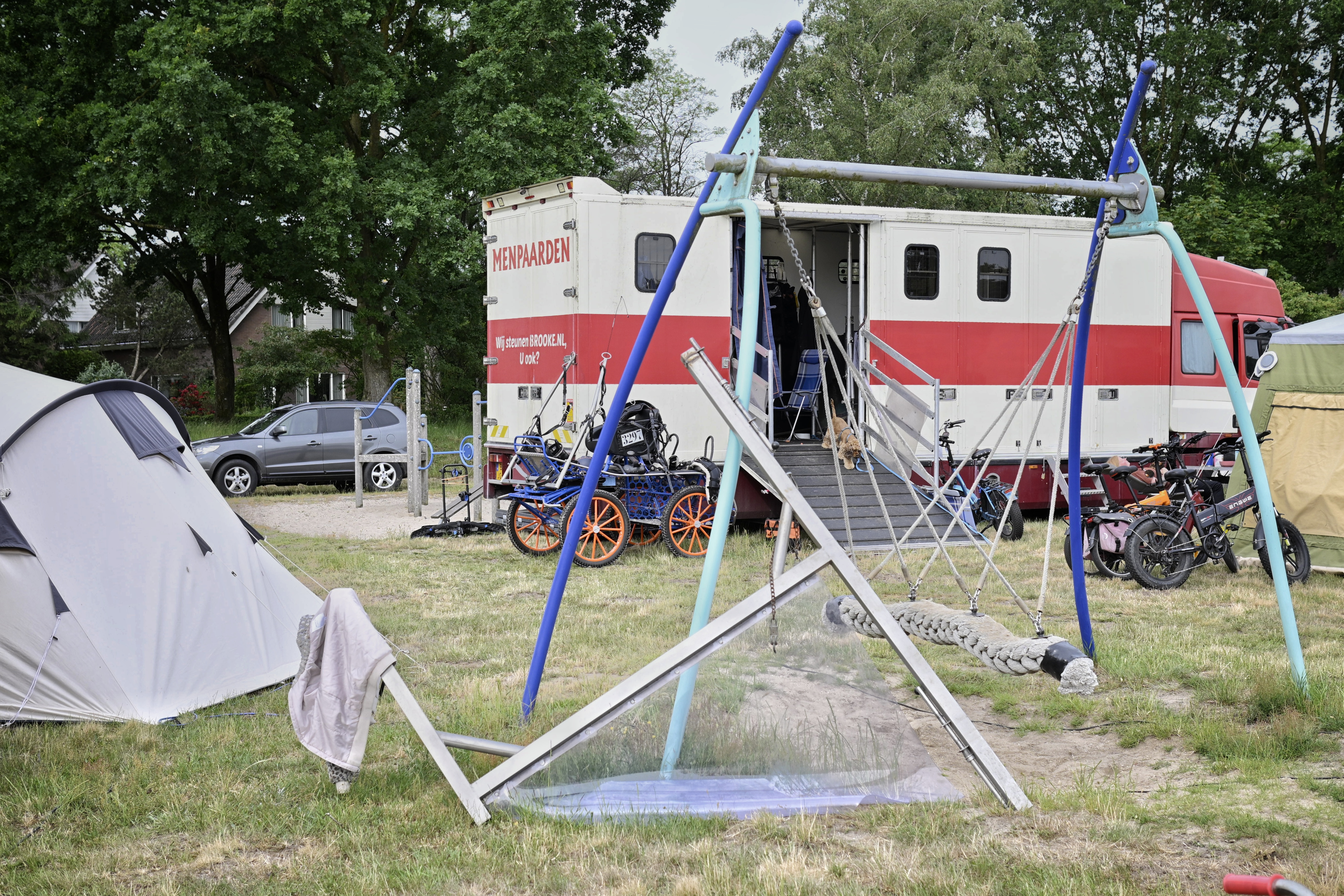 A steel construction leans against a swingset. A tent, a trailer and a horse cart surround it.