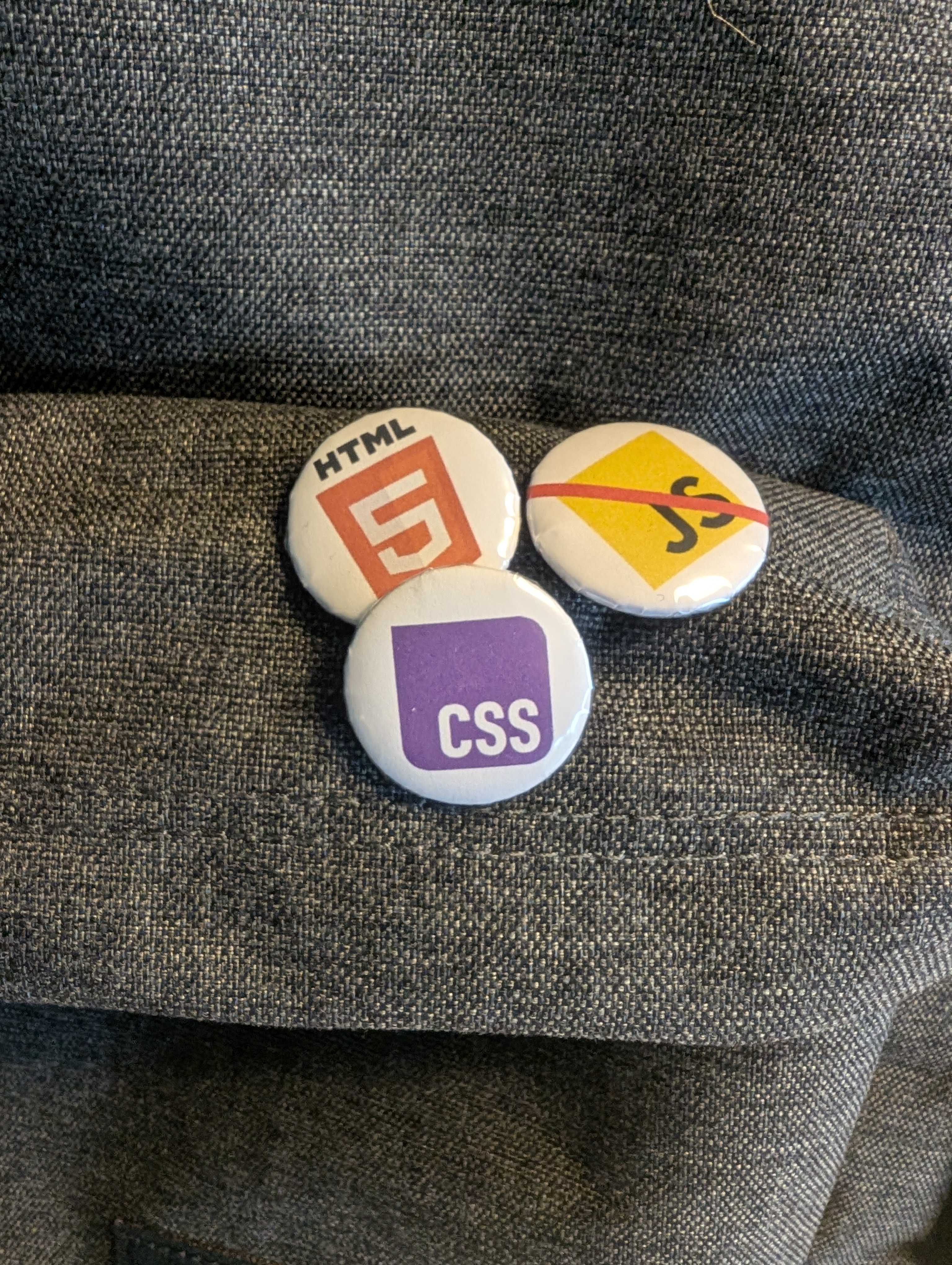 Three buttons on dark fabric. One with an HTML-logo, another with a CSS-logo, and the third has a JS-logo but it's crossed out.